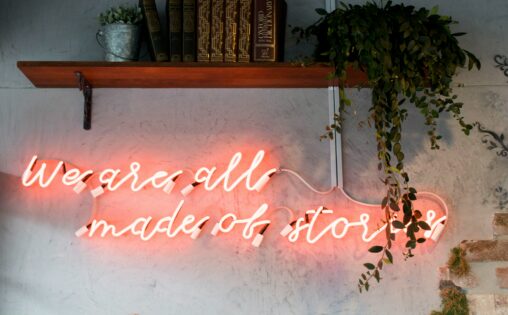 Neon cursive writing that says "We are all made of stories" under a wooden wall shelf with books and a plant.