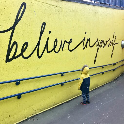 A child in a yellow coat looking up at a yellow wall painted with the message "believe in yourself"